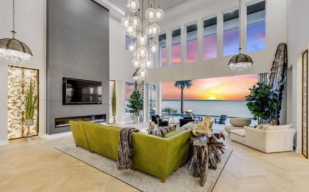 Double-height living room with fireplace wall, chandelier cluster, and sunset water view.